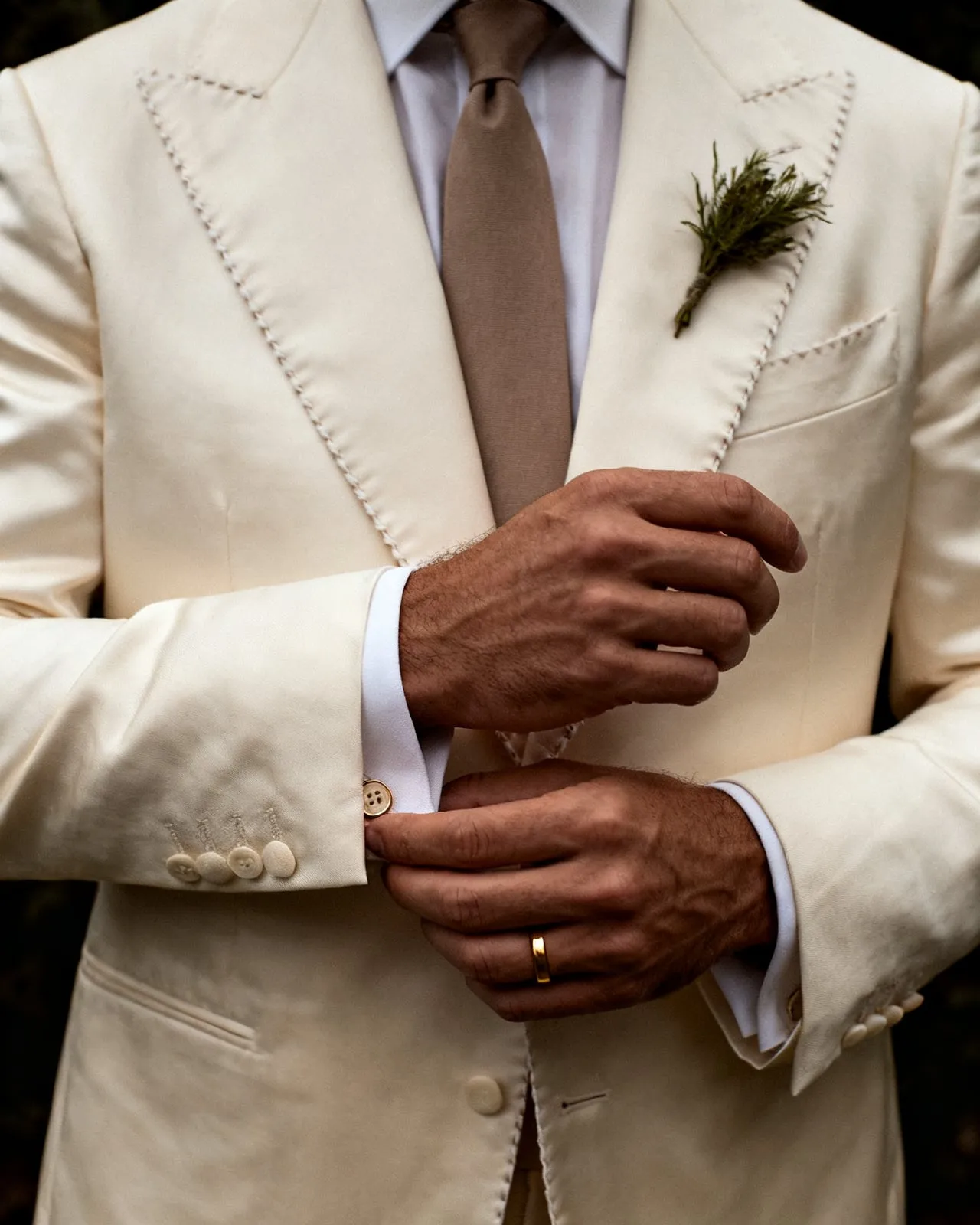 A groom's hands fastening the cuff of a hand-stitched cream silk jacket, a gold band on his finger and a sprig of rosemary at the buttonhole