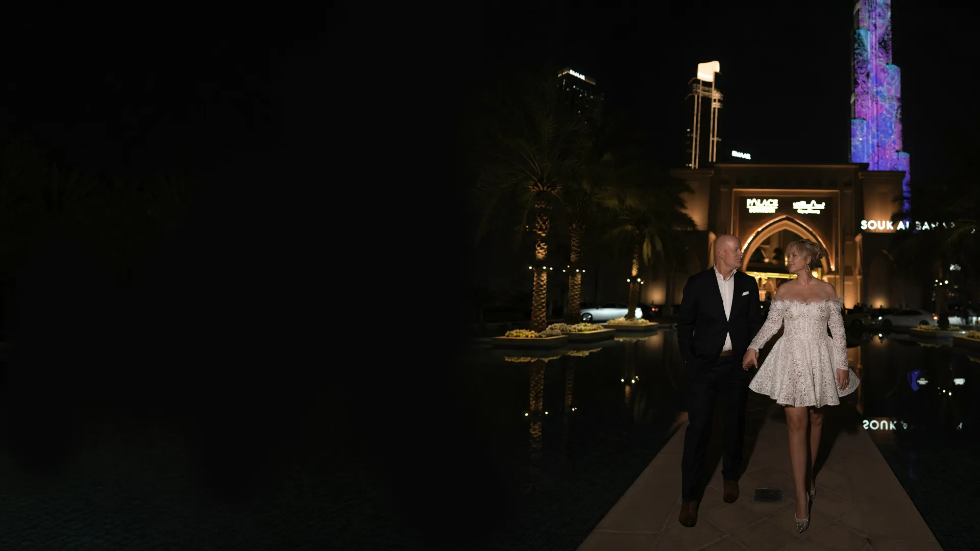 A couple in evening wedding attire walking hand in hand beside a lit lagoon in Downtown Dubai at night, the Burj Khalifa rising behind them