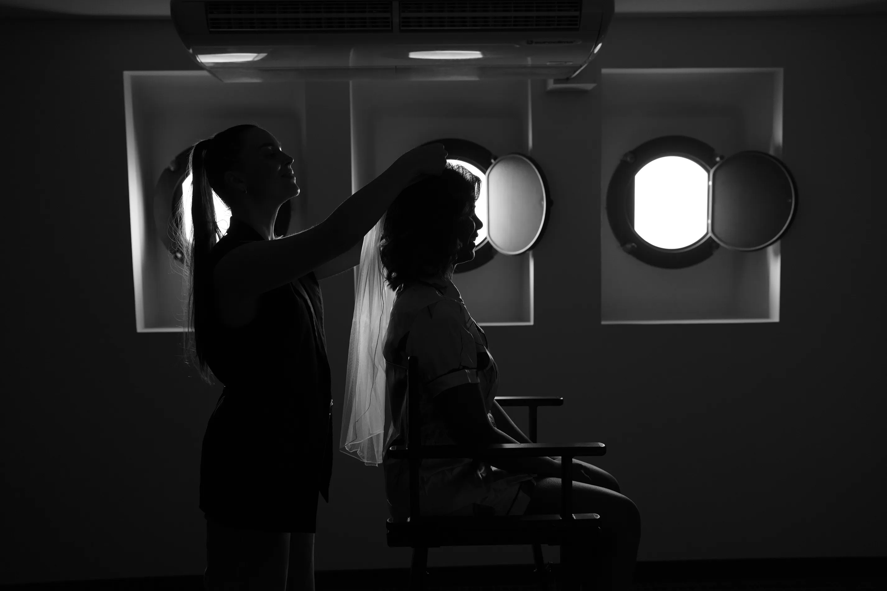 A black-and-white frame of a stylist lifting a bride's veil into place as she sits in a director's chair in front of two studio softboxes